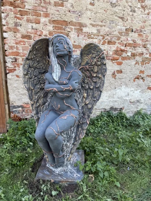 Duża grafitowo-miedziana figura ogrodowa przedstawiająca siedzącą postać anioła z zamkniętymi oczami, skrzyżowanymi rękami i dużymi dekoracyjnymi skrzydłami.