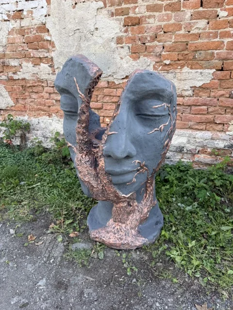 Duża czarno-miedziana figura ogrodowa przedstawiająca stylizowaną twarz rozdartą na pół, z dekoracyjnym motywem pnia i gałęzi drzewa biegnącym przez środek rzeźby.
