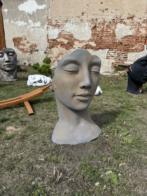 Szara figura ogrodowa przedstawiająca stylizowaną twarz kobiety z zamkniętymi oczami i dekoracyjną, rzeźbiarską formą.