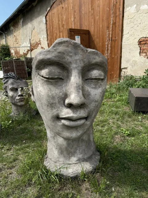Duża szaro-brązowa figura ogrodowa o wysokości 114 cm, przedstawiająca stylizowaną twarz kobiety z zamkniętymi oczami oraz praktycznym siedziskiem umieszczonym z tyłu.