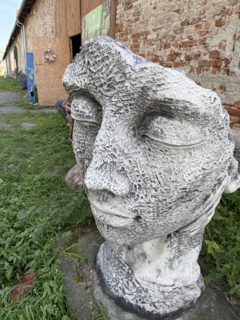 Duża biało-szara figura ogrodowa przedstawiająca stylizowaną twarz z zamkniętymi oczami, spokojnym wyrazem oraz wyraźnie zaznaczoną fakturą powierzchni.