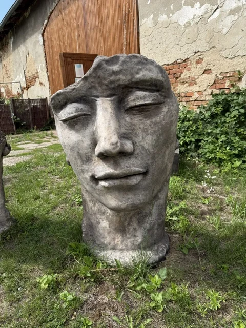 Duża szaro-brązowa figura ogrodowa o wysokości 114 cm, przedstawiająca stylizowaną twarz mężczyzny o wyrazistych rysach, z praktycznym siedziskiem umieszczonym z tyłu.